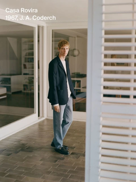 Young man in a navy blazer and light-wash jeans stands on a tiled porch outside a modern house with white-painted shutters and trim.