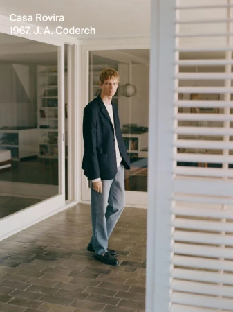 Young man in a navy blazer and light-wash jeans stands on a tiled porch outside a modern house with white-painted shutters and trim.