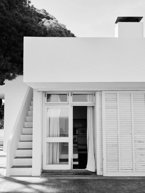 Black and white architectural shot of a modern white building with stairs, window, door, and a shuttered window, showcasing a minimalist aesthetic.