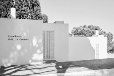 Monochrome architectural photograph of the exterior of Casa Rovira featuring white plastered walls and tree shadows.