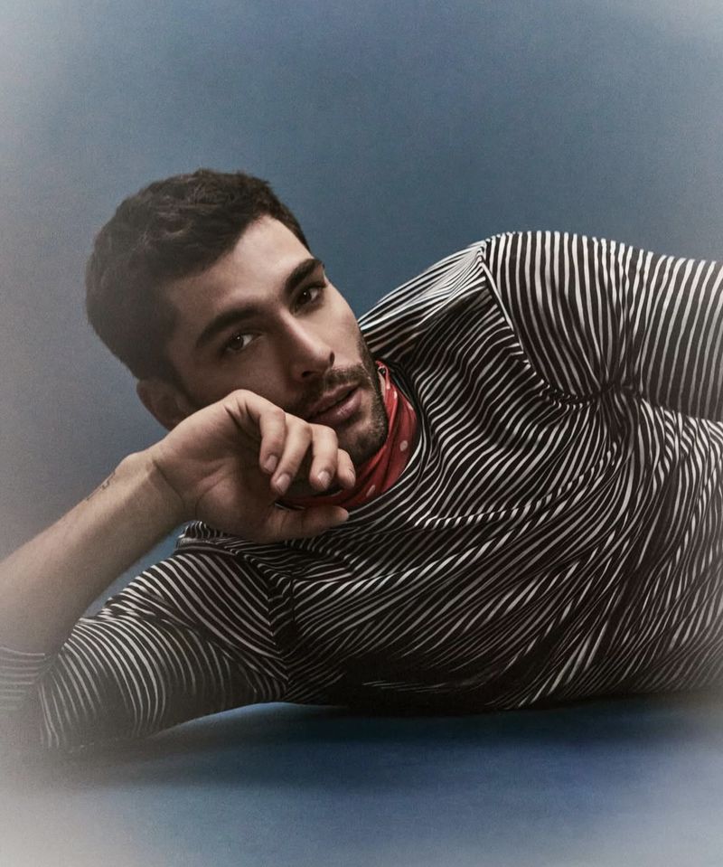 A male model with dark hair is lying down wearing a striped shirt and a red bandana, posing in a studio with a blue backdrop.