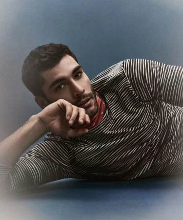 A male model with dark hair is lying down wearing a striped shirt and a red bandana, posing in a studio with a blue backdrop.