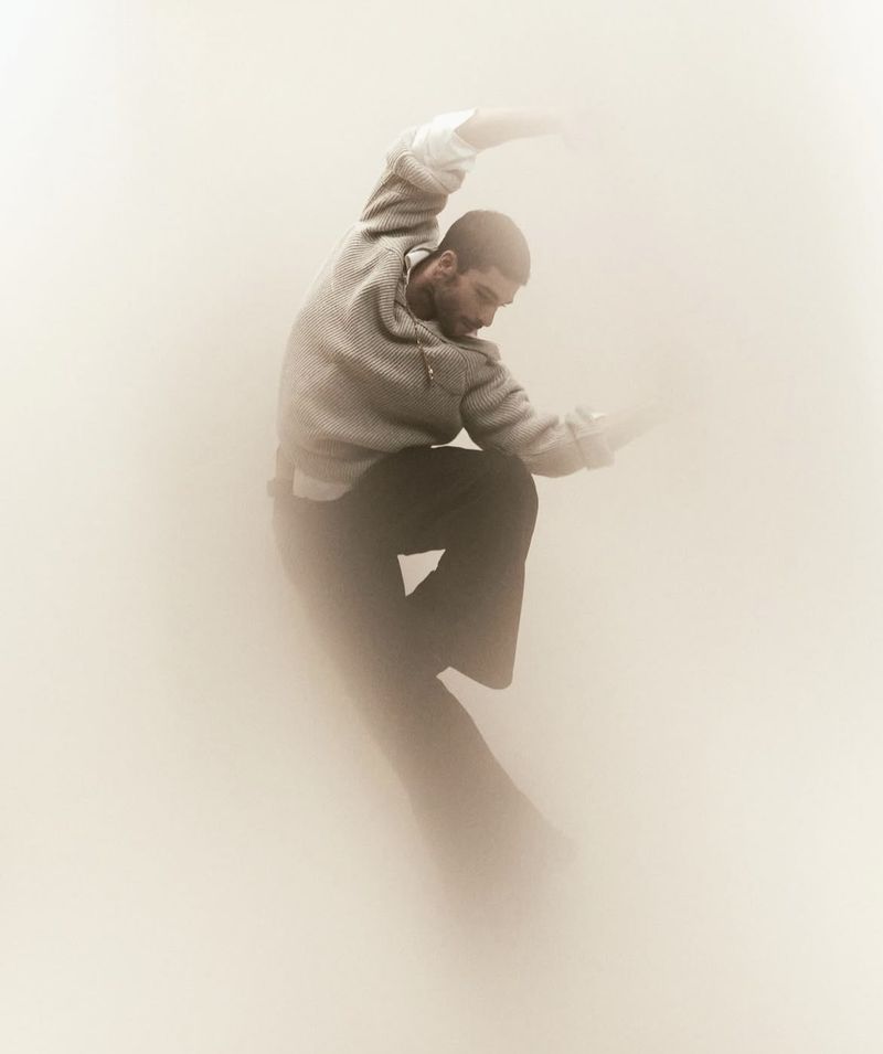 An adult male with a beard is depicted in a dynamic pose against a soft, diffused background, wearing a grey top and dark pants.