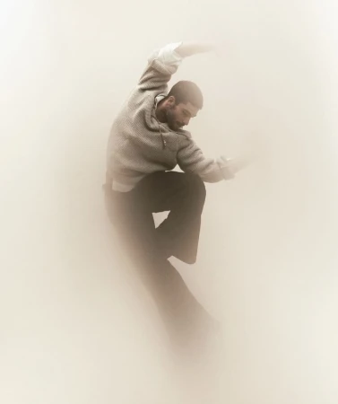 An adult male with a beard is depicted in a dynamic pose against a soft, diffused background, wearing a grey top and dark pants.