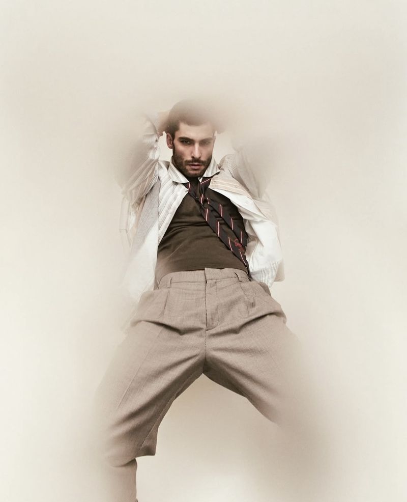 Male fashion model poses in an open white shirt, brown vest, and tie, against a light backdrop, demonstrating a unique and artistic style.