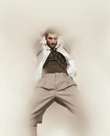 Male fashion model poses in an open white shirt, brown vest, and tie, against a light backdrop, demonstrating a unique and artistic style.