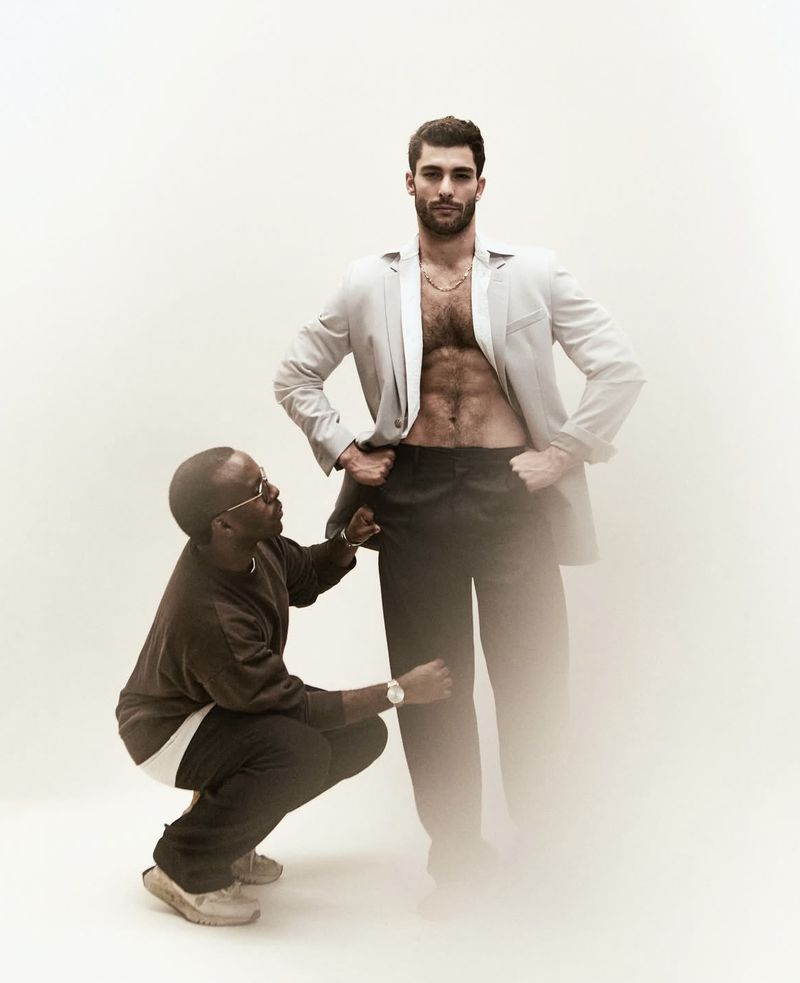 Attentive tailor measuring a muscular man's trousers. The man is wearing a jacket, no shirt, and dark pants in a bright studio.
