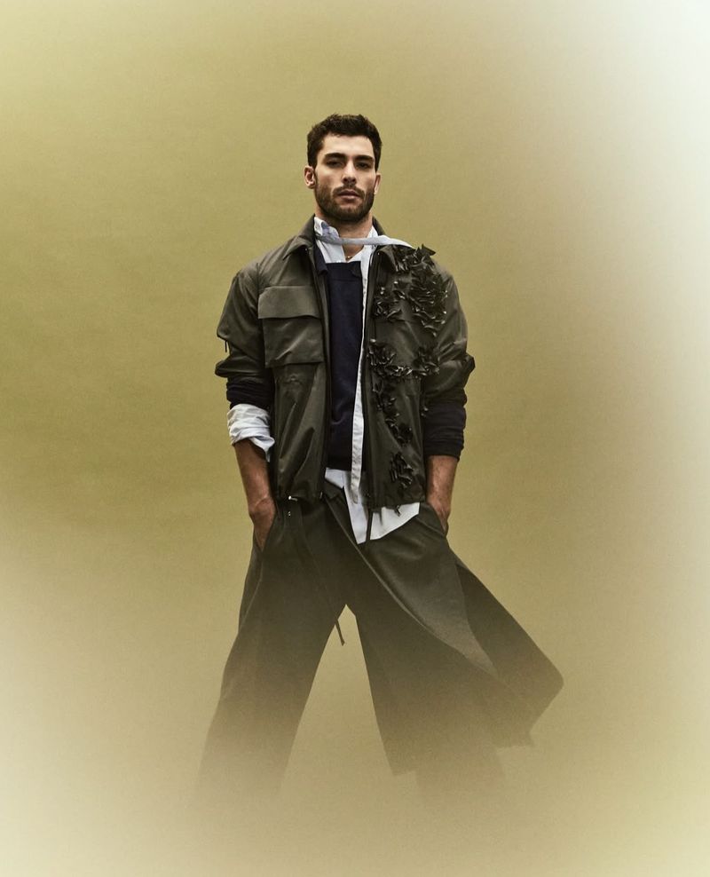 Portrait of a stylish male model in a green jacket and wide leg pants against a neutral background in a fashion editorial photo.