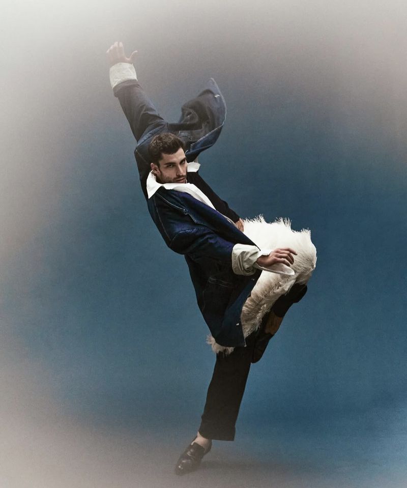 A male dancer wearing a denim jacket and dark trousers performs a dynamic pose against a blue background in a fashion editorial style.