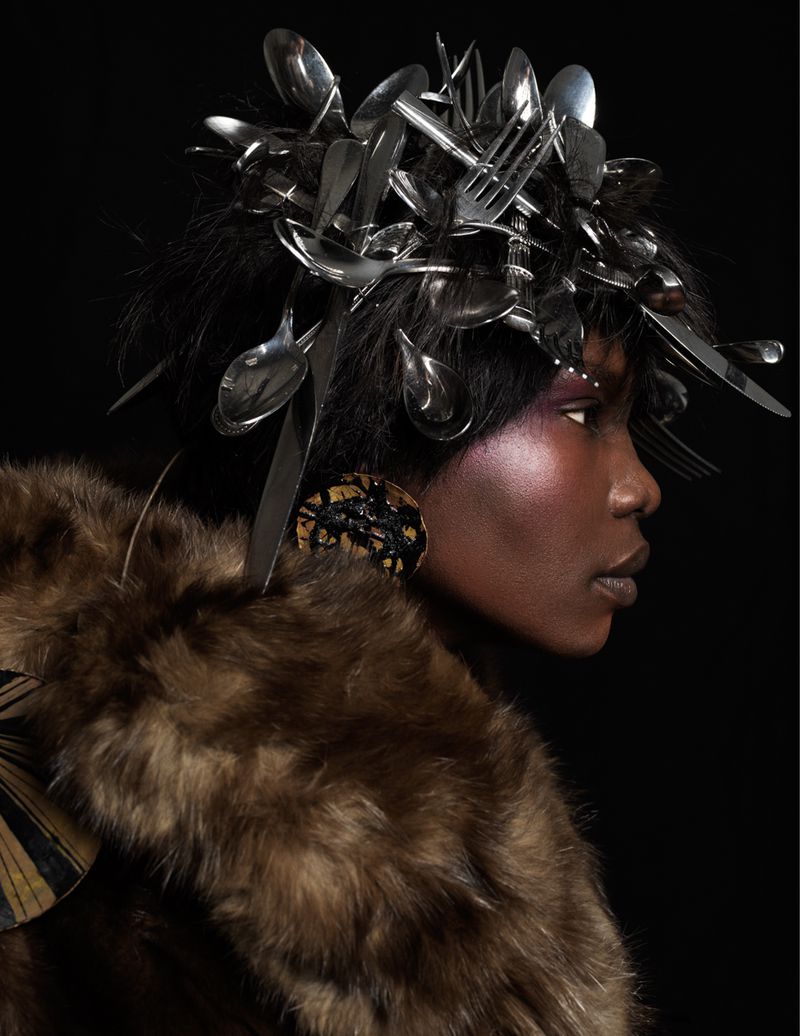 Fashion shot of an African American woman with a silverware headpiece and a fur coat against a dark background in a studio.
