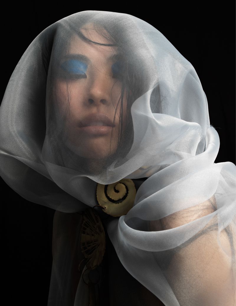 Woman with blue eyeshadow wearing a translucent white veil with decorative shell pendants against a dark backdrop in a portrait.