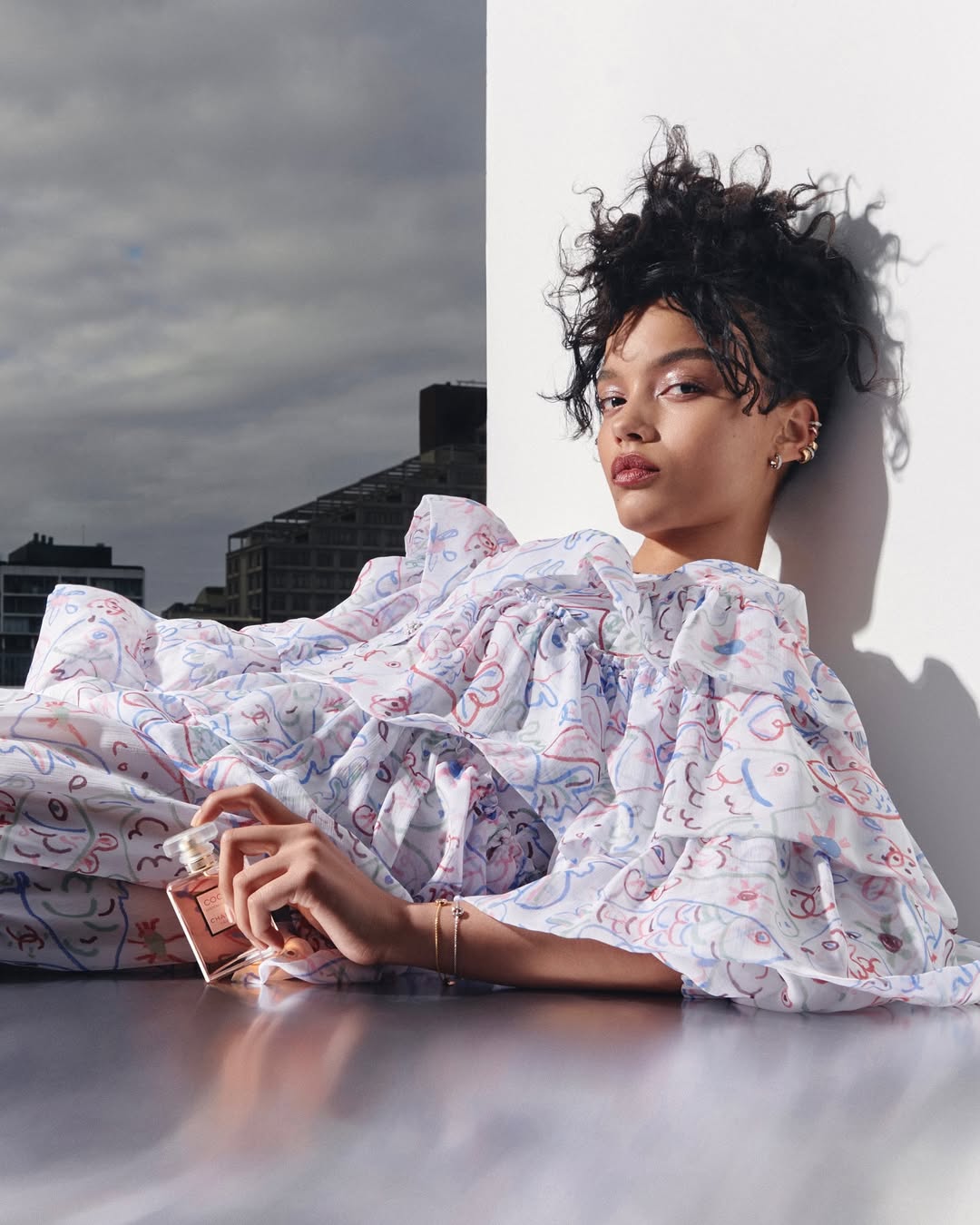 Elegant woman in a ruffled dress holds perfume bottle against a split backdrop of city buildings and white wall in a beauty shot.