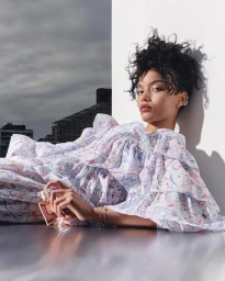 Whitney Peak leaning against a white wall holding a perfume bottle wearing a patterned dress
