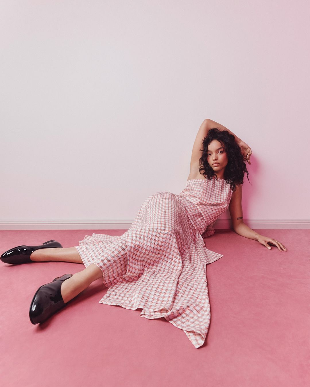 A woman in a pink checkered dress poses on a pink floor in front of a cream-colored wall in a fashion studio shot.