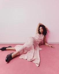 Whitney Peak reclining on a pink carpet wearing a checkered pink dress and black loafers against a matching wall