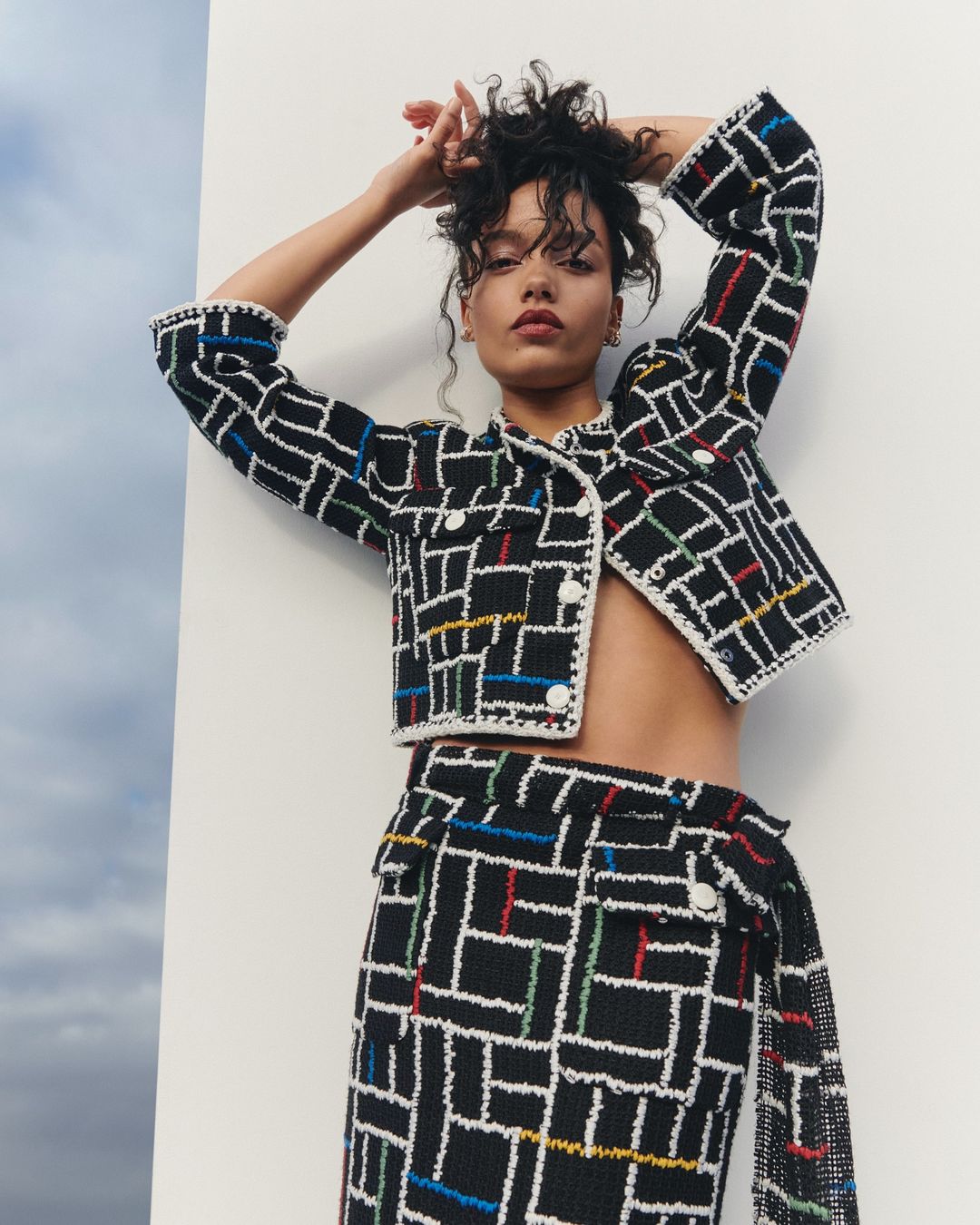 A curly-haired woman in a fashionable black and white tweed jacket and skirt set poses outdoors against a cloudy sky and white wall.
