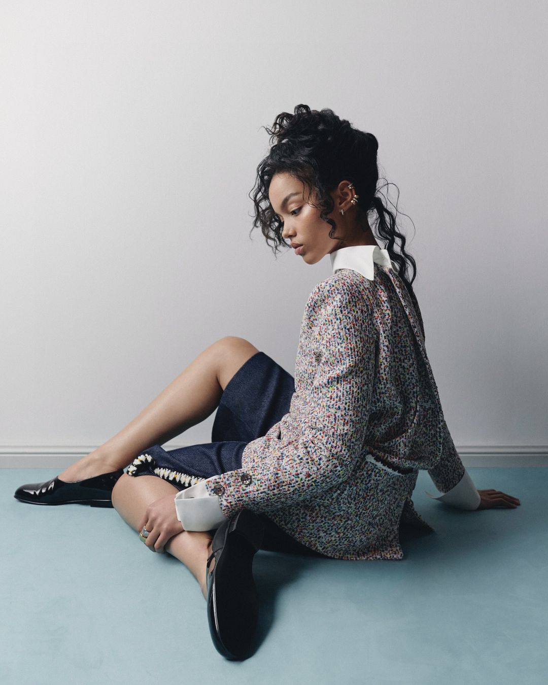 Portrait of a woman sitting on the floor, wearing a patterned blazer, denim skirt, and black shoes in a studio setting.