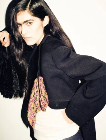 A woman with long dark hair poses indoors, wearing a black blazer, white top, and blue jeans, with a floral purse on her shoulder.