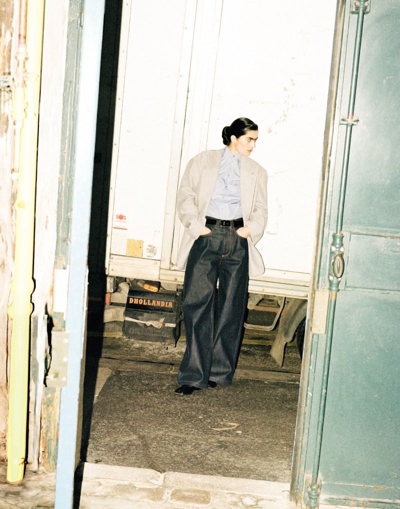 A fashion shot presents a person in a blazer and wide-leg jeans standing near a doorway with a stack of boxes and a white door.