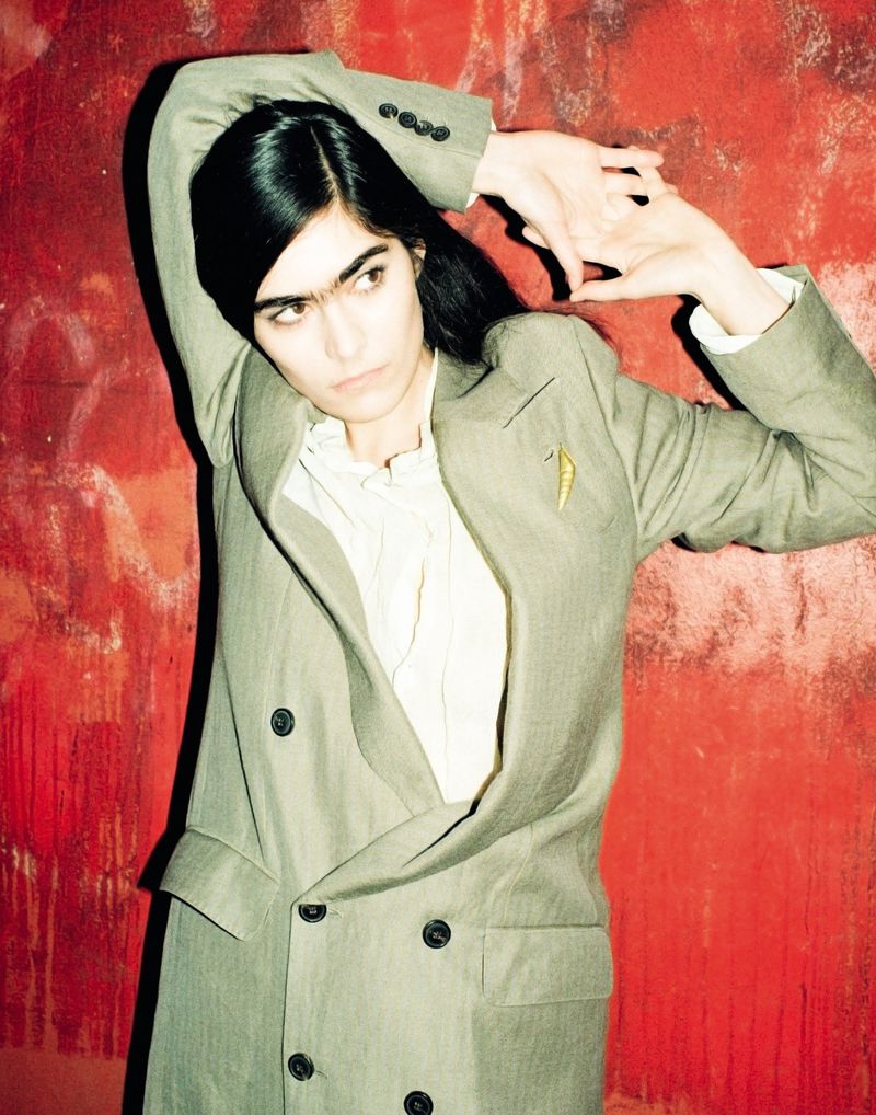 Woman in a grey double-breasted suit jacket and a white shirt, standing against a textured red background and raising arms above head.