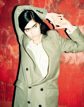 Woman in a grey double-breasted suit jacket and a white shirt, standing against a textured red background and raising arms above head.