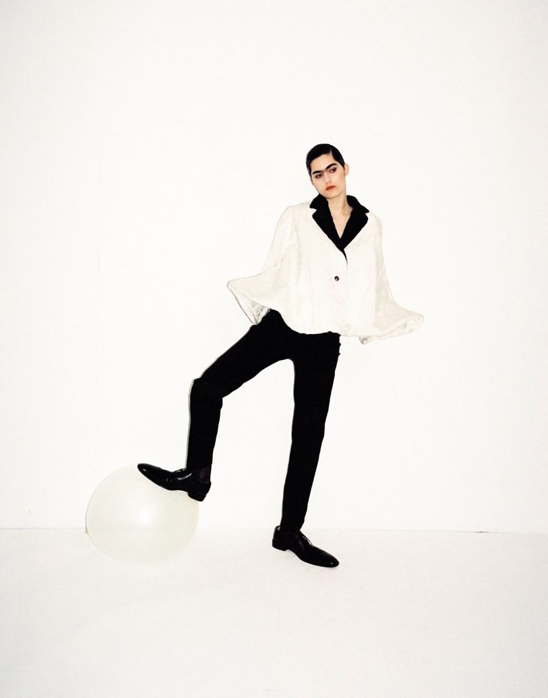Fashionable young adult poses with a balloon in a studio, wearing a white jacket with black collar, black pants, and black shoes.