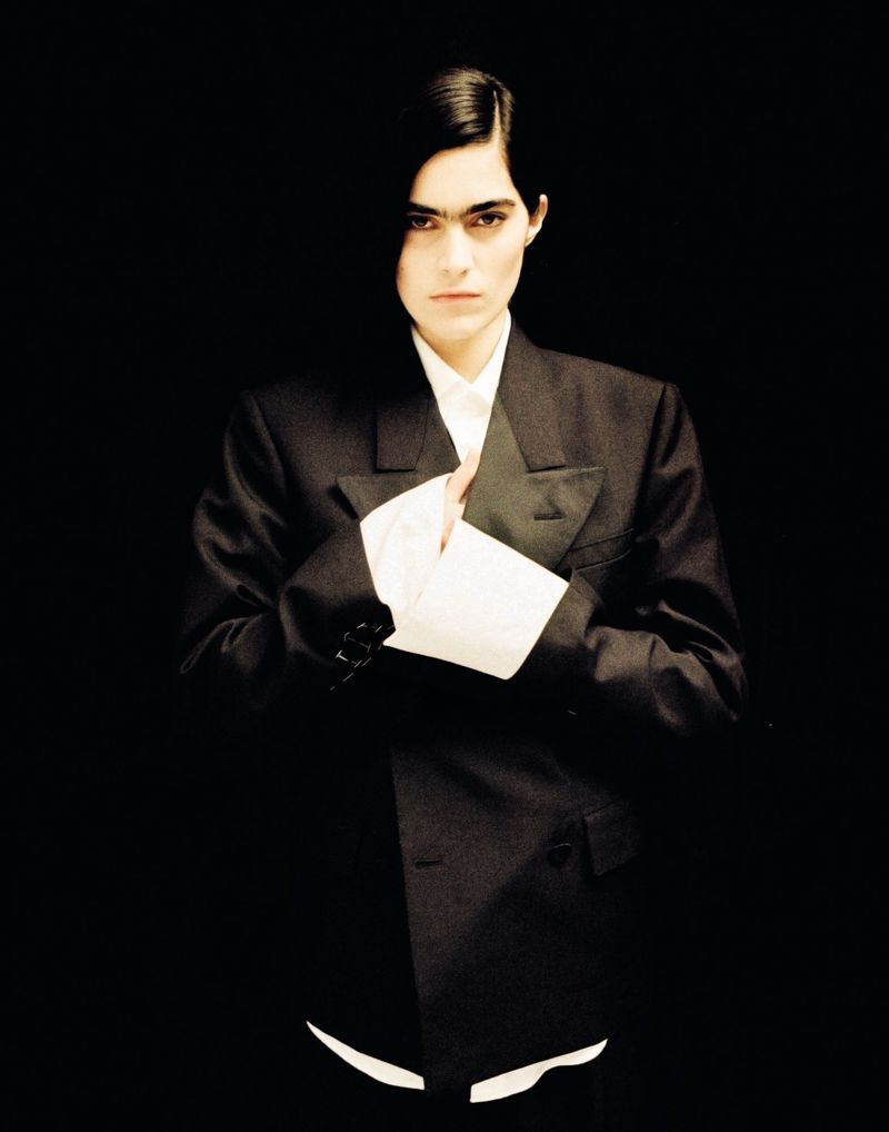 Portrait of a woman in a dark suit with a white collared shirt against a dark backdrop, emphasizing fashion and style.