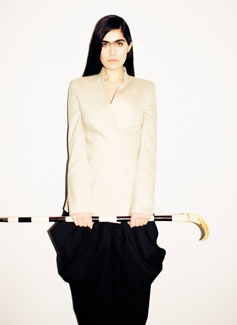 Woman in cream jacket and black skirt holds striped cane, looking at the camera against a white backdrop in a studio setting.