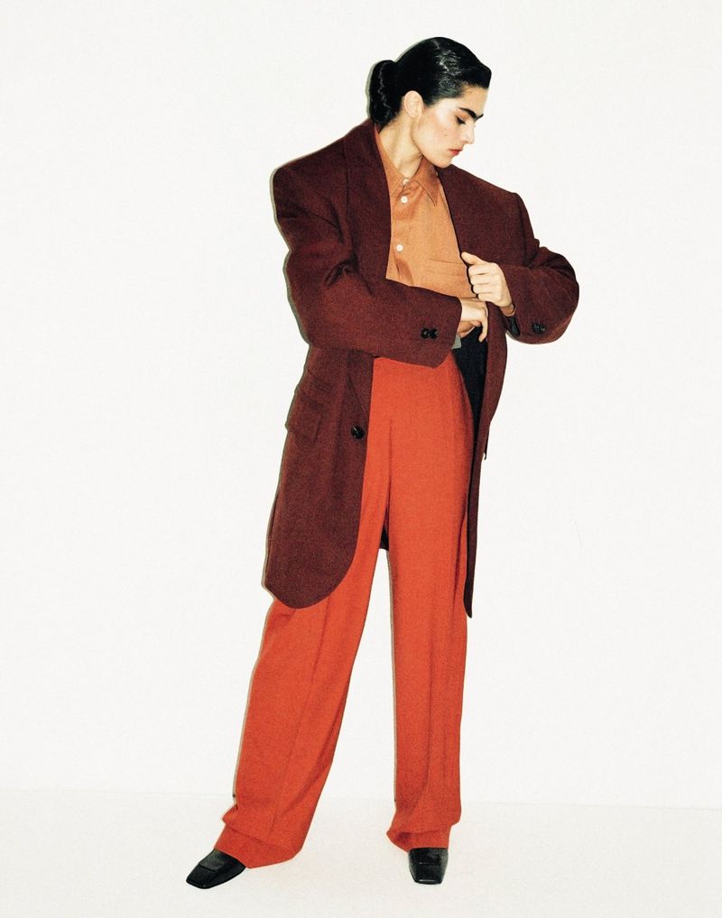 Adult fashion model poses in a full-length shot wearing an auburn overcoat, copper button-down shirt, and red-orange slacks against a white background.