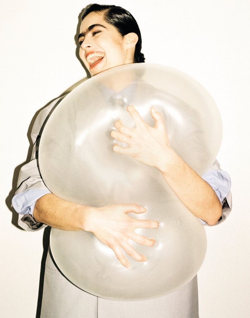 Portrait of a smiling adult with a wet hair look hugging a large transparent balloon in a photo studio with a neutral backdrop.