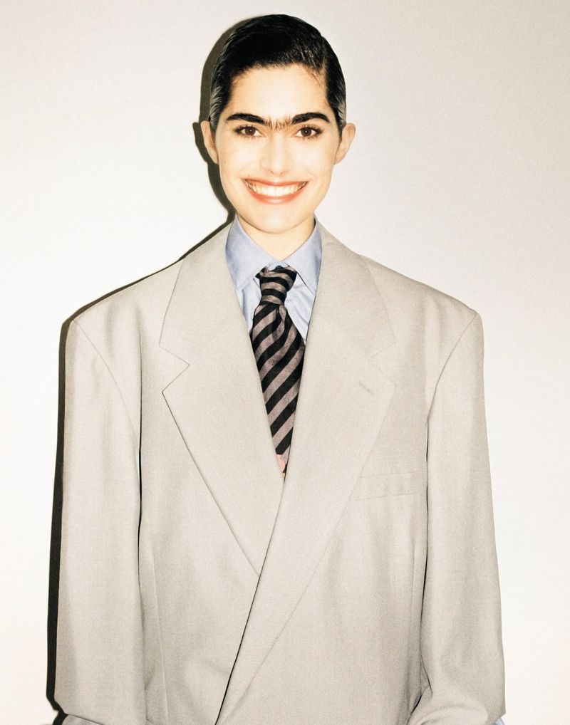 Smiling woman in oversized grey business suit with a striped tie stands indoors in front of a neutral white wall.