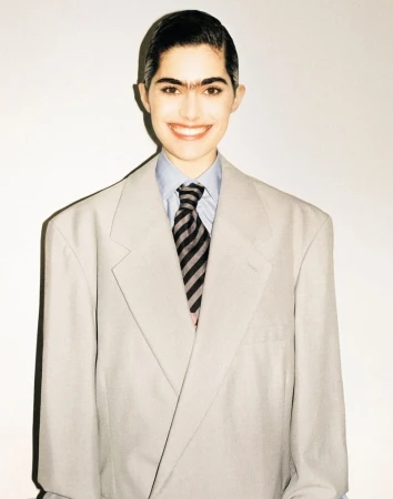 Smiling woman in oversized grey business suit with a striped tie stands indoors in front of a neutral white wall.