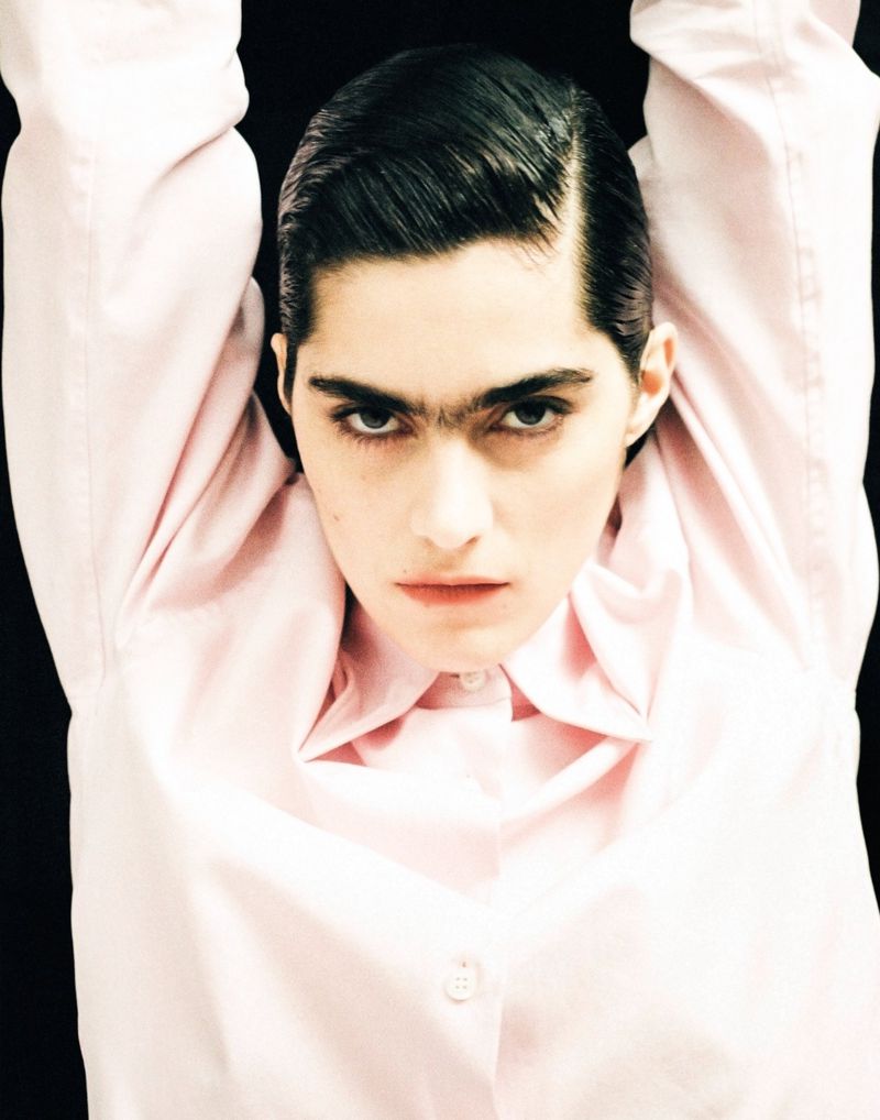 A striking portrait of a woman with unique brows, wearing a light pink collared shirt, set against a stark black background.
