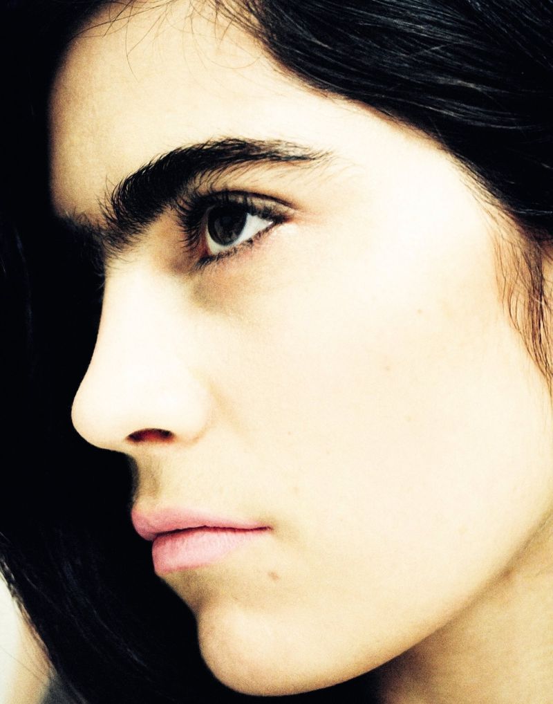 Close-up portrait of an adult woman with dark hair looking to the side, set against a dark background with filtered yellow and white light.