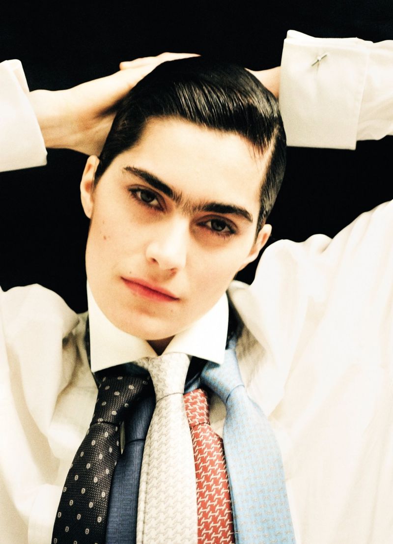 Androgynous person in a white shirt and multiple ties poses with hands behind their head against a stark black background.