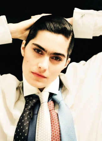 Androgynous person in a white shirt and multiple ties poses with hands behind their head against a stark black background.