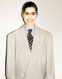 Model in an oversized grey suit jacket, light blue shirt, and striped tie against a plain white studio backdrop.
