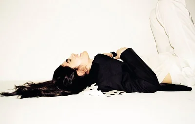 A laughing woman with long dark hair lying on her back against a white background wearing a black jacket and white trousers.