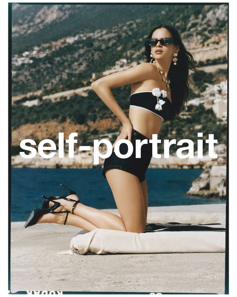 Woman in black and white bikini and high heels poses on a rooftop with a sea and mountain backdrop, fashion photography.