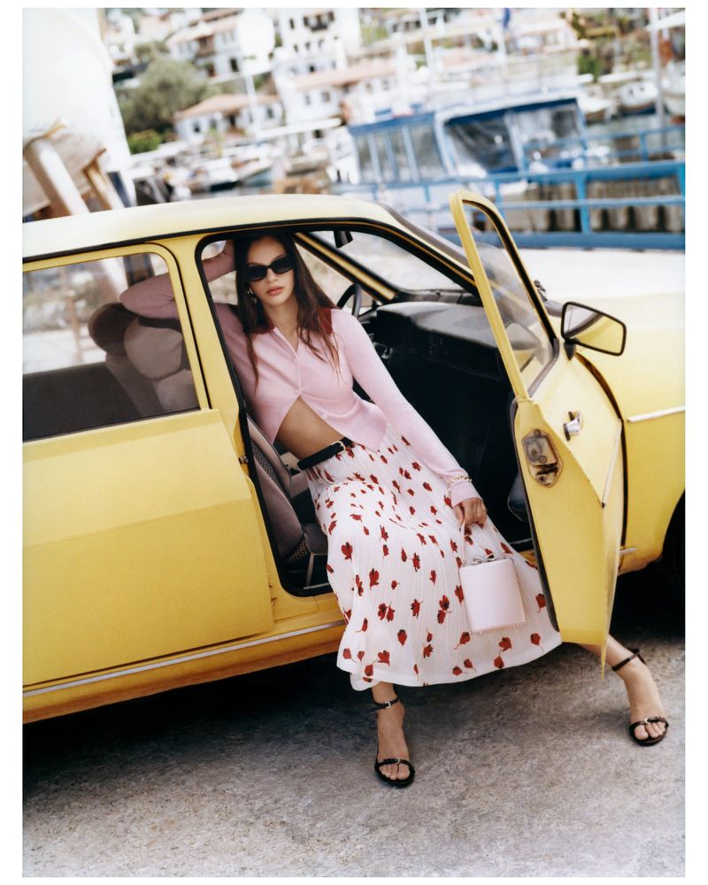 A young adult woman in sunglasses wearing a skirt with red floral print poses next to a vintage yellow car with a coastal background.