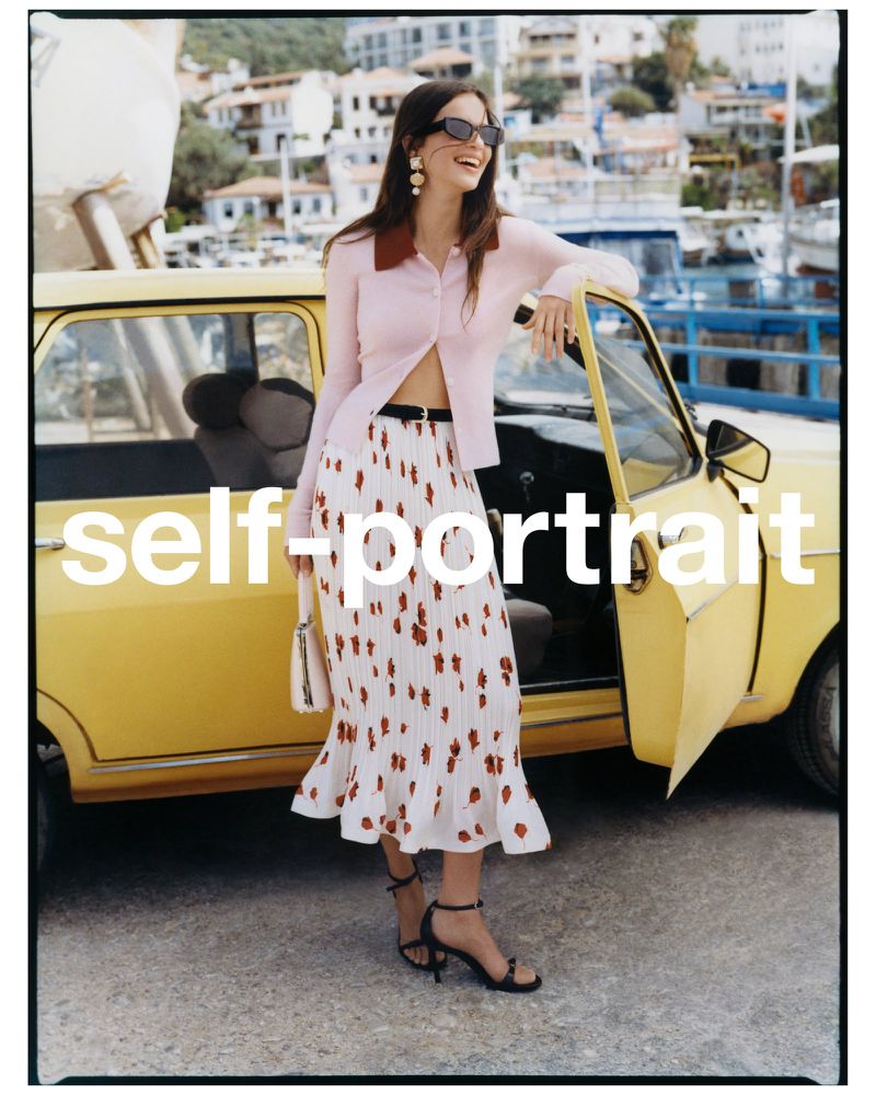 A woman poses in a light pink collared shirt and patterned skirt with a vintage yellow car in an outdoor setting with buildings and boats.