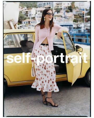 A woman poses in a light pink collared shirt and patterned skirt with a vintage yellow car in an outdoor setting with buildings and boats.