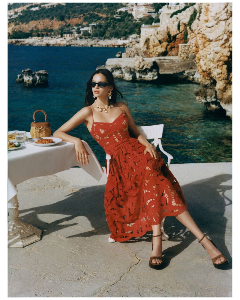 Woman in red dress poses at table with ocean view. Summer fashion shoot featuring architecture and natural coastal scenery.