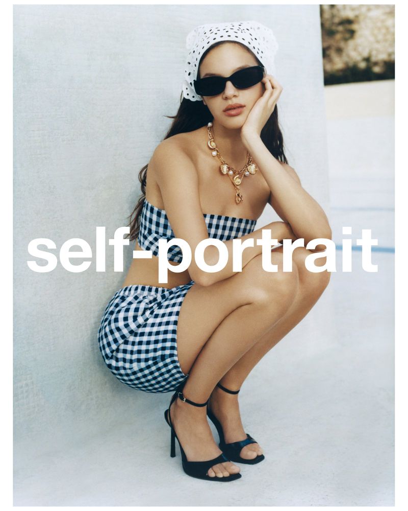 A woman in a gingham two-piece set and black high heels poses against a light background, accessorized with a headscarf and jewelry.