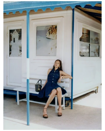Young woman in a denim dress sits on a white bench outside a beach house, holding a black purse. She wears platform heels.