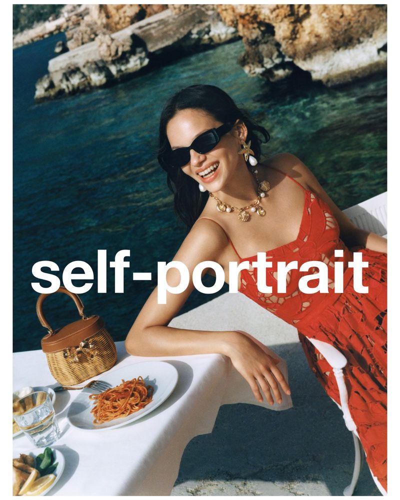 Model in red dress wearing sunglasses and statement necklace leaning on a table near the coast for Self-Portrait PF '25.