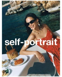 Model in red dress wearing sunglasses and statement necklace leaning on a table near the coast for Self-Portrait PF '25.