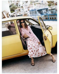 Model sitting in a vintage yellow car wearing a pink cardigan and white patterned skirt holding a handbag