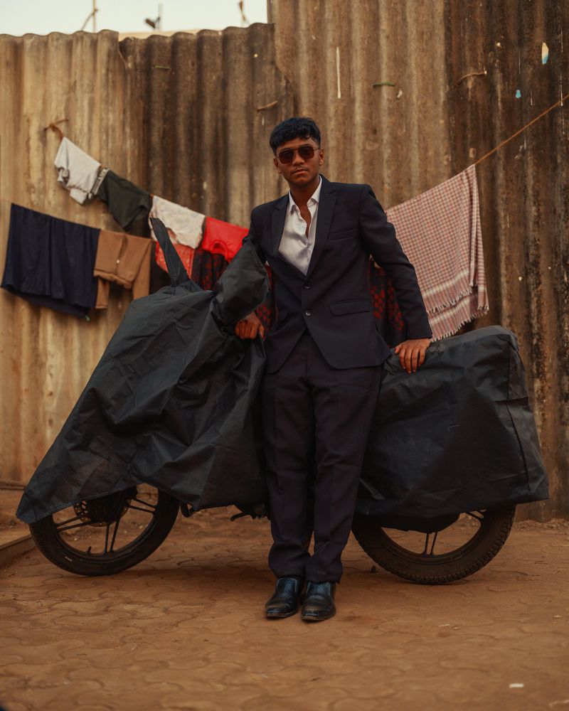 Man in suit poses next to a motorcycle covered by a tarp, against a corrugated metal wall with hanging laundry in outdoor setting.
