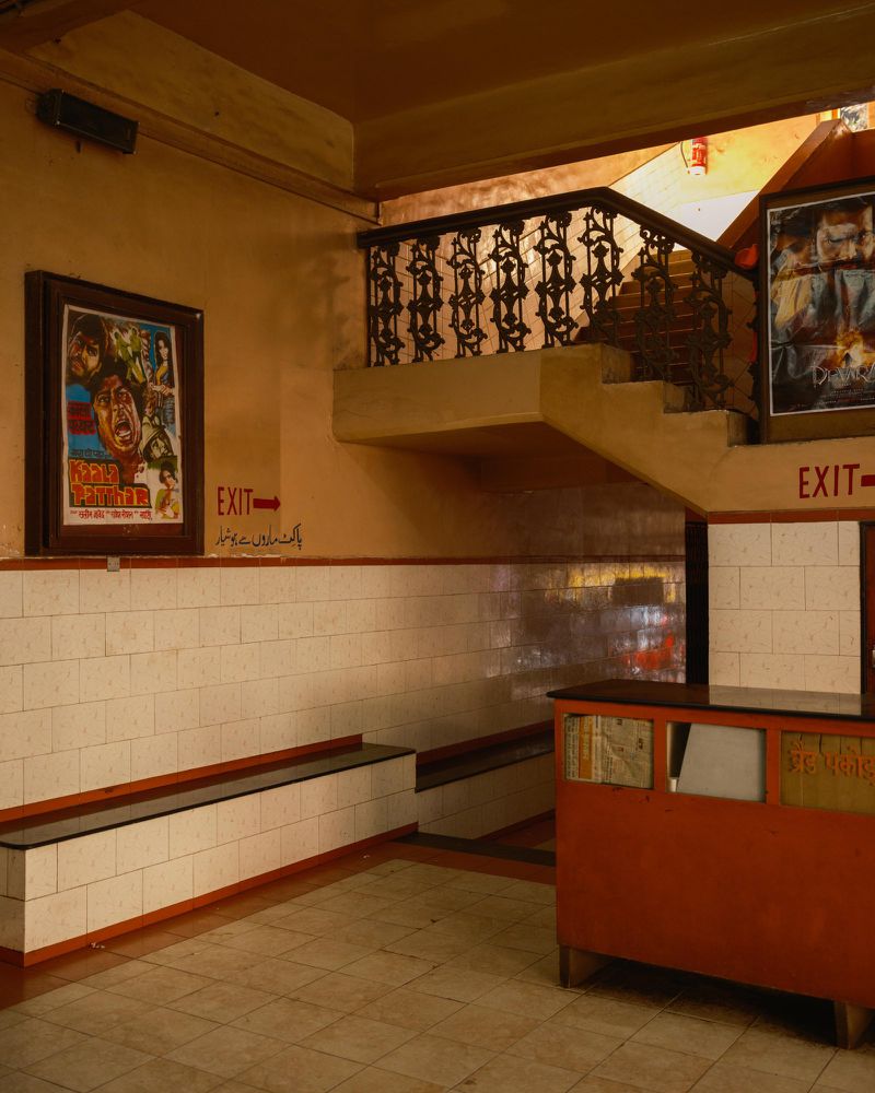 Indoor scene of a building lobby with a staircase, walls with white tiles, posters, and an exit sign with orange and beige color tones.